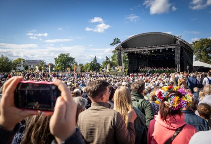 Na pierwszym planie telefon, którym nagrywany jest koncert, w tle publiczność wraz z olbrzymią sceną na której są Artyści