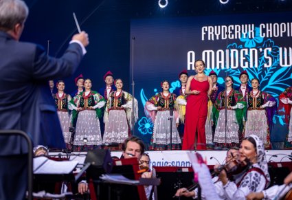 W lewym roku Dyrektor Jacek Boniecki zwrócony w stronę orkiestry i Artystów na scenie. Na środku sceny Solistka w długiej, czerwonej sukience a za nią Artyści w kostiumach krakowskich