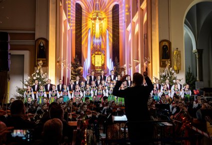 Na pierwszym planie postać Dyrygenta zwrócona w stronę Orkiestry w kostiumach wilanowskich i Artystów Chóru w kostiumach łowickich