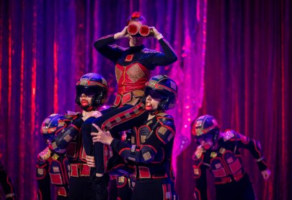 Scena postaci kwadratów. Postać kobieca patrzy przez czerwono-czarną lornetkę, siedząc na barkach innych postaci kwadratów. Postacie męskie mają kaski