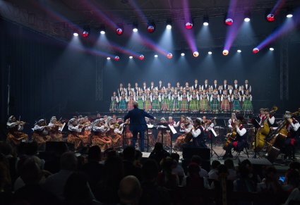 Na podświetlonej scenie znajdują się Artyści Chóru w kostiumach łowickich w trakcie śpiewu, niżej Artyści Orkiestry w kostiumach wilanowskich oraz Dyrygent zwrócony w ich stronę