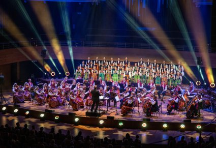 Widok na scenę na której znajduje się Dyrygent zwrócony w stronę Orkiestry w kostiumach wilanowskich i Artystów Chóru w kostiumach łowickich