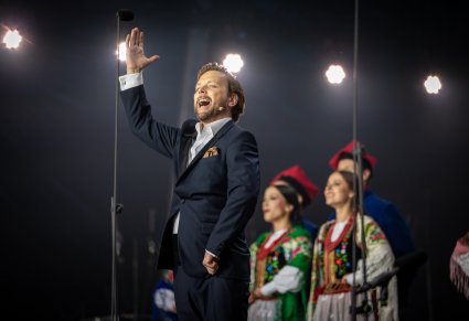 Zbliżenie na śpiewającego Solistę w garniturze a za nim część Artystów w kostiumach krakowskich