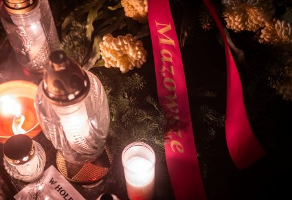Zbliżenie na wiązankę z ozdobną taśmą "Mazowsze", obok znicze, zdjęcie wykonane wieczorem