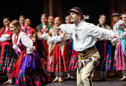 Zbliżenie na parę Artystów w kostiumach podhalańskich w trakcie wykonywania choreografii tanecznej. W tle reszta część Artystów również w kostiumach podhalańskich