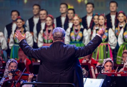 Zbliżenie na Dyrektora Jacka Bonieckiego, który jest zwrócony w stronę Orkiestry, za orkiestrą Chór w kostiumach łowickich