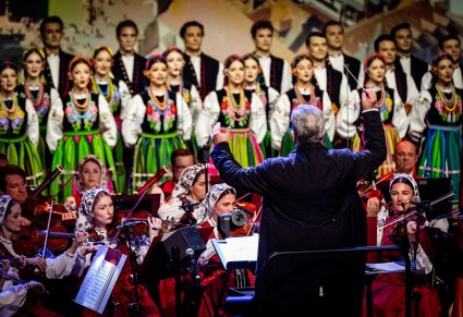 Dyrektor Jacek Boniecki zwrócony w stronę Orkiestry, za orkiestrą widoczni Artyści z chóru w kostiumach łowickich ustawieni w rzędy