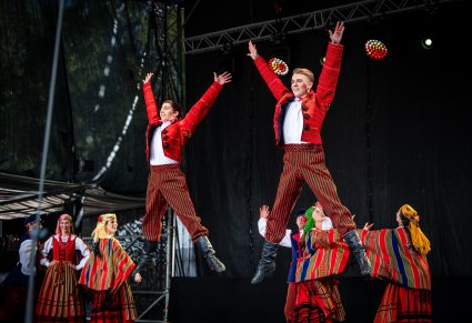 Dwóch Artystów w kostiumach sieradzkich na pierwszym planie w figurach tanecznych a na drugim planie reszta Artystów