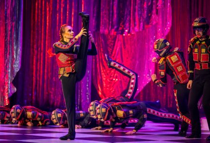 Na pierwszym planie postać kobieca grająca postać kwadratu. Postać wysoko podnosi nogę.