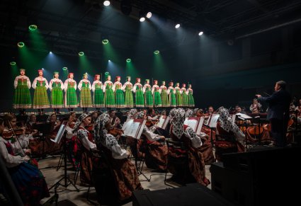 Na pierwszym planie orkiestra w kostiumach wilanowskich z Dyrygentem, na scenie Artyści chóru w kostiumach kurpiowskich w trakcie śpiewu