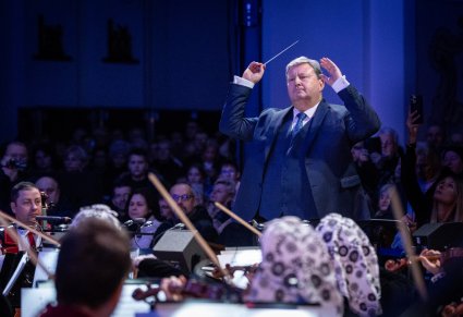 Zbliżenie na dyrygującego Dyrektora zespołu "Mazowsze", przed nim Orkiestra w kostiumach wilanowskich