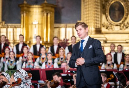 Zbliżenie na Solistę w garniturze, na drugim planie widoczna część Chóru w kostiumach łowickich