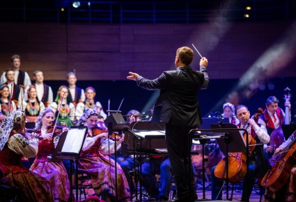 Na pierwszym planie Dyrygent z batutą zwrócony w stronę Orkiestry w kostiumach wilanowskich, widoczna część Artystów Chóru w kostiumach łowickich