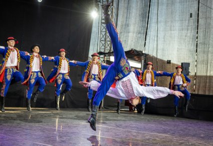 Na środku sceny Artysta Baletu w trakcie wykonywania akroobacji tanecznych, za nim Artyści w kostiumach podegrodzkich wykonujący choreografię taneczną