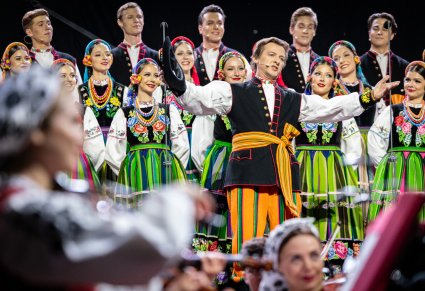 Na pierwszym planie artysta chóru, wykonujący "Furmana", za nim część chóru, wszyscy w kostiumach łowickich, poniżej część orkiestry w kostiumach wilanowskich