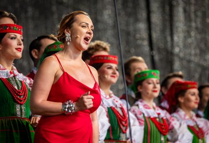 Solistka w czerwonej sukni obok Artystów chóru, którzy mają na sobie kostiumy kurpiowskie