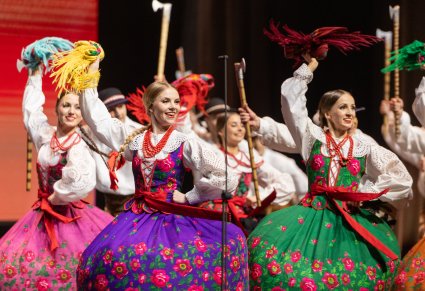 Na pierwszym planie grupa Artystek w trakcie wykonywania choreografii tanecznej, Artystki machają w górze kolorowymi chustami