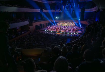 Widok na salę Narodowego Forum Muzyki, na scenie Artyści Chóru w kostiumach łowickich, Orkiestra w kostiumach wilanowskich wraz z Dyrygentem. Widoczna publiczność.