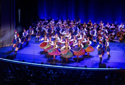 Na scenie Artyści w kostiumach krakowskich podczas wykonywania choreografii tanecznej, w tle widoczna orkiestra