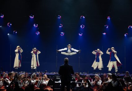 Na scenie Artyści baletu w kostiumach łańcuckich podczas wykonywania imponującej choreografii tanecznej, widoczny Dyrygent zwrócony w stronę Orkiestry  sceny