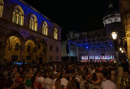 Widok na publiczność pod gołym niebem, publiczność znajduje się  na placu między budynkami a przed nią scena z Artystami