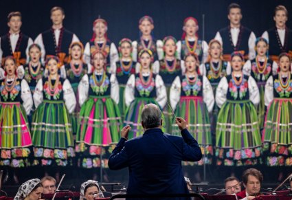 Na pierwszym planie Dyrygent trzymający w ręku batutę, zwrócony w stronę Artystów Orkiestry w kostiumach wilanowskich oraz Artystów Chóru w kostiumach łowickich