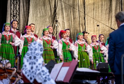 Chór na scenie w kostiumach kurpiowskich z widoczną orkiestrą i Dyrygentem