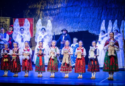 Przedszkolaki w kostiumach ludowych trzymające w rękach pluszowe maskotki, za nimi część postaci ze spektaklu