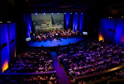 Widok na publiczność i scenę z góry. Na scenie Dyrektor Jacek Boniecki, Orkiestra i Artyści chóru w kostiumach łowickich