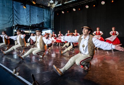 Na pierwszym planie mężczyźni wykonujący spektakularne figury z układu tanecznego, w tle kobiety wykonujące choreografię taneczną. Artyści w kostiumach jurgowskich