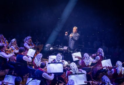 Na pierwszym planie Dyrektor zespołu "Mazowsze" trzymający w ręku batutę i dyrygujący Orkiestrze w kostiumach wilanowskich