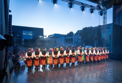Widok na Artystów chóru w kostiumach łowickich stojących na scenie, zwróconych w stronę Publiczności.