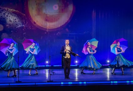 Na scenie Solista w smokingu, trzymający kolorowy parasol i grupa Tancerek wykonująca choreografię taneczną
