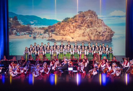 Na pierwszym planie Dyrektor Jacek Boniecki dyrygujący koncert, Orkiestra a za nią chór w kostiumach łowickich ustawiony w rzędy