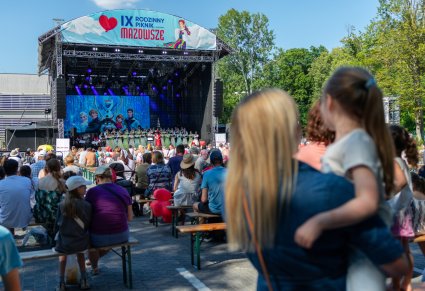 Na pierwszym planie kobieta trzymająca na ręku dziecko, obydwie są zwrócone w stronę sceny głównej na której znajdują się Artyści Chóru i Orkiestry