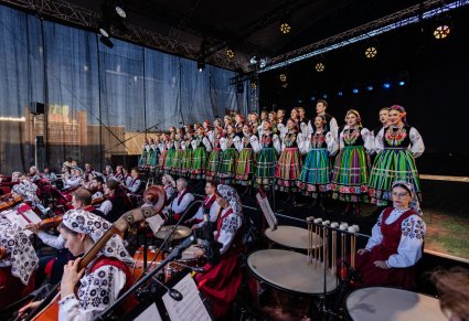 Boczny widok na Artystów chóru w kostiumach łowickich, stojących w dwurzędzie, niżej sceny Artyści Orkiestry w kostiumach wilanowskich, grający na instrumentach