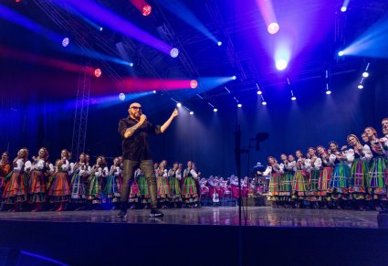Na środku sceny Solista zespołu Kombii w trakcie śpiewu, po jego bokach Artyści zespołu "Mazowsze" w kostiumach ludowych, a w tle sceny Orkiestra w kostiumach wilanowskich wraz z Dyrygentem