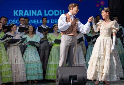Na pierwszym planie para Artystów śpiewająca partię Solistyczne w kostiumach stylizowanych XIX wiecznych, w tle część Artystów Chóru również w takich kostiumach