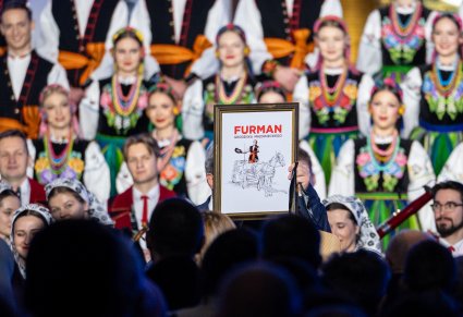 Na pierwszym planie obraz w ramce przedstawiający zabawną grafikę "Furman Grodziska Mazowieckiego", w tle część Artystów Chóru w kostiumach łowickich