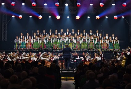 Na scenie Artyści Chóru w kostiumach łowickich, niżej Artyści Orkiestry w kostiumach wilanowskich wraz z Dyrygentem, widoczna część Publiczności