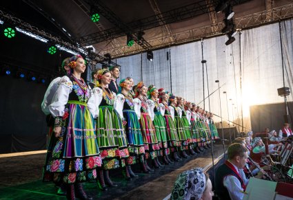 Na scenie Artyści chóru w kostiumach łowickich, stojący w dwurzędzie, niżej sceny widoczna część Orkiestry w kostiumach wilanowskich