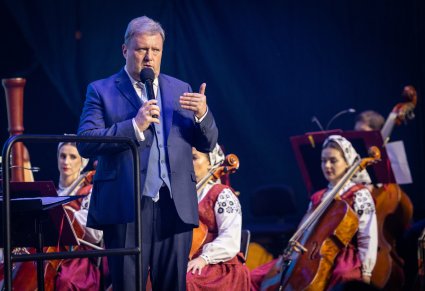 Zbliżenie na Dyrektora zespołu "Mazowsze" wypowiadającego się do mikrofonu. W tle część Artystów orkiestry w kostiumach wilanowskich