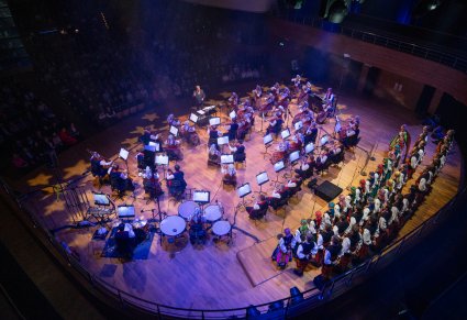 Widok z góry na scenę na której znajduje się Chóru zespołu w kostiumach łowickich ustawiony w 3 rzędach. Przed nim Orkiestra w kostiumach wilanowskich wraz z Dyrygentem z Batutą, w tym przypadku jest to Dyrektor zespołu "Mazowsze"