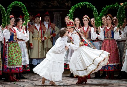 Panna Młoda w kostiumie krakowskim z czepcem złotogłowym wykonująca choreografię taneczną wraz z Artystką w zwiewnym, białym kostiumie, w tle widoczna część Artystów w kostiumach krakowskich