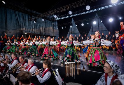 Na scenie Artyści w kostiumach ludowych w trakcie ukłonów, niżej część orkiestry