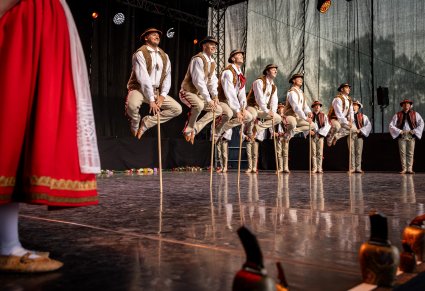 Grupa Artystów na środku sceny w kostiumach jurgowskich wykonująca choreografie taneczną z drewnianymi kijkami