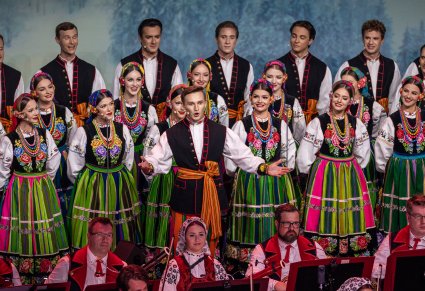 Zbliżenie na Solistę a za nim część Artystów chóru w kostiumach łowickich ustawionych w rzędzie. Na dole zdjęcia widoczne 4 osoby z Orkiestry