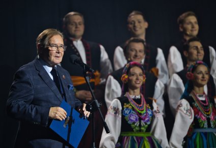 Zbliżenie na prowadzącego koncert, za nim widoczna część Chórzystów w kostiumach łowickich