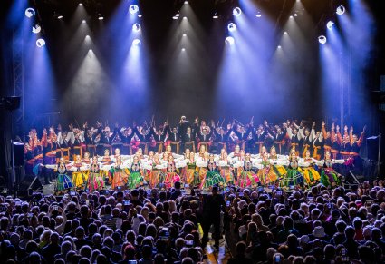Widok na scenę i Publiczność. Na scenie Artyści zespołu "Mazowsze" w kostiumach ludowych podczas wykonywania finałowej choreografii, w tle sceny Orkiestra w kostiumach wilanowskich