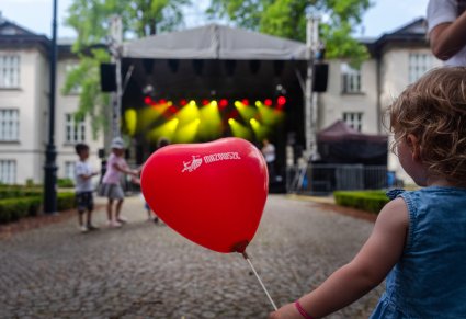Na pierwszym planie czerwony balon w kształcie serca z logo zespołu "Mazowsze" trzymany w rączce małej dziewczynki, w tle Mała Scena plenerowa